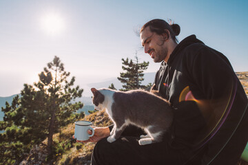 Male tourist and his cat with a mug of water on top of a mountain at sunrise