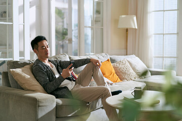 young asian man sitting in family couch in living room at home looking at mobile phone - obrazy, fototapety, plakaty