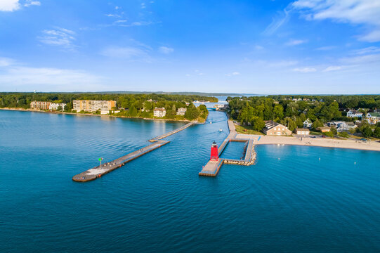 Aerial drone photograph of serene river inlet from Lake Michigan into Charlevoix harbor under a vivid afternoon blue sky, reflecting on gently rippled turquoise water bordered by lush green shorelines