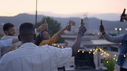 Diverse people toasting with alcoholic beverages at barbecue party on rooftop in the evening. Multi-ethnic friends clinking beer bottles on terrace. Smiling men and women having fun together outdoors