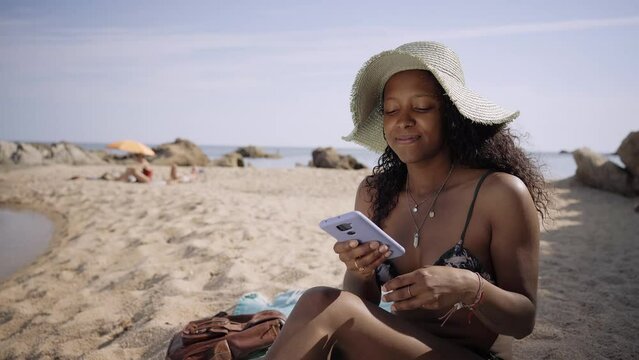 Attractive young Latin girl sitting on beach talking on phone. Female tourist in swimsuit and straw hat African American woman enjoying summer vacation on Spanish coast. People enjoying good weather.