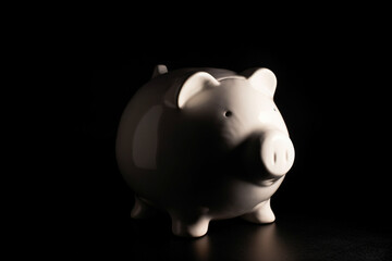 A white porcelain piggy bank filled to the brim with silver coins, illuminated by a single spot light, against a dark background.
