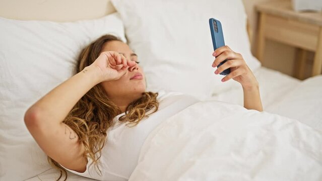 Young beautiful hispanic woman using smartphone tired lying on bed at bedroom