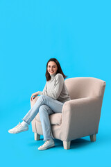 Obraz premium Young woman sitting in soft armchair on blue background