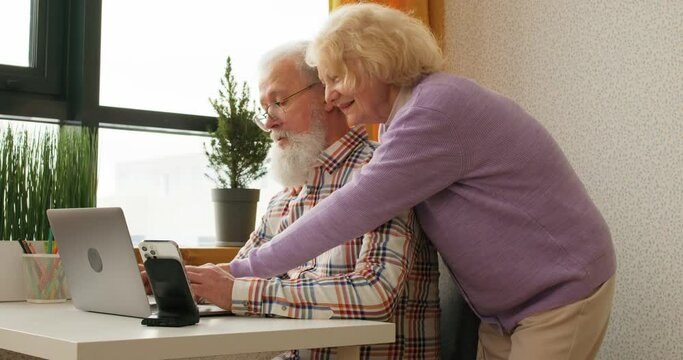 Happy smiling elderly mature couple having fun online, surfing the Net. active social networks users spend time together, watching something interesting, grandpa and grandma talking laughing happiness