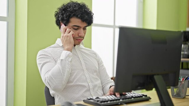Young latin man business worker using computer talking on smartphone at office