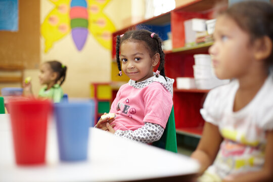 Child, classroom and lunch in portrait with smile for education at a table with children. Break, kids and learning at school with apple in kindergarten with chair or desk with food with friends.
