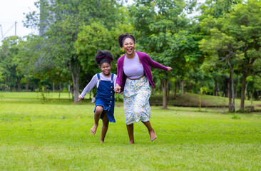Naklejka na ściany i meble African American mother running barefoot on the grass lawn with her young daughter while having a summer picnic in the public park for wellbeing and happiness