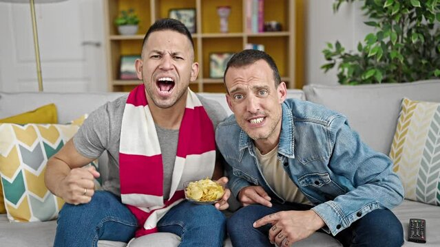 Two men supporting soccer team sitting on sofa at home