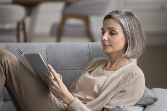 Focused pretty grey haired woman reading bestseller paper book at home, enjoying literature, leisure, relaxation, studying new knowledge, relaxing on comfortable sofa at home