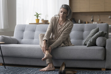 Frustrated concerned mature woman taking high heeled shoes off, sitting on home sofa, touching...