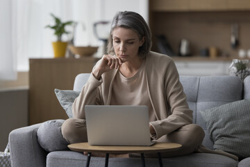 Serious thoughtful mature freelancer woman working at laptop, sitting on couch at home, looking at display, thinking on freelance job project, online education, browsing website, watching content