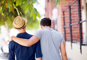 City, travel and back of gay couple walking together in the street while on a tourism vacation. Love, affection and male friends embracing in an outdoor urban road while on a weekend trip or holiday.
