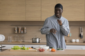 Cheerful African American chef man in cook hat dancing to music at kitchen table with salad ingredients, enjoying cooking, homemade culinary activities, smiling, laughing, cutting vegetables