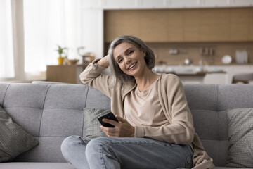 Obraz premium Happy mature naturally grey haired woman holding mobile phone, resting on sofa, looking at camera, posing indoors, smiling, laughing, using digital gadget for domestic Internet communication