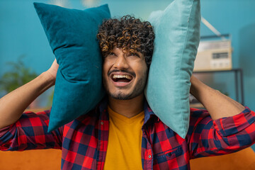 Repair work at neighbours. Irritated indian man relaxing on couch cover ears with pillows annoyed...