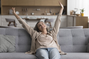Positive awaking mature woman sitting on comfortable sofa, stretching body, rising hands up,...