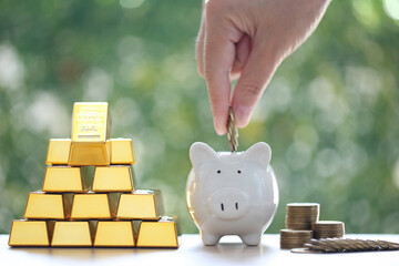 Woman hand putting a coin into piggy bank and gold bar on natural green background,Business...