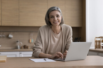 Cheerful positive mature freelance business woman educating for new job, working at laptop computer in kitchen, typing, looking at camera, smiling. Home female portrait