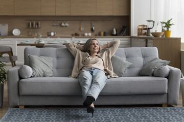 Obraz premium Happy dreamy calm grey haired woman resting on comfortable sofa, enjoying lazy weekend, leisure at modern cozy home, breathing cool air, looking away, thinking, daydreaming