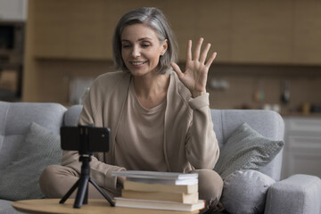 Positive grey haired teacher woman speaking at educating online conference, recommending learning books for studying, education, waving greeting hand at smartphone screen, talking on video call