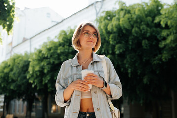 Portrait of cute smiling young woman wearing glasses and casual clothes with cup of coffee in park looking away, summer lifestyle