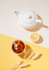 Creative layout of a glass cup of herbal tea with lemon, flowers and fruits and teapot on a white and yellow background with a shadow. Healthy drink concept.
