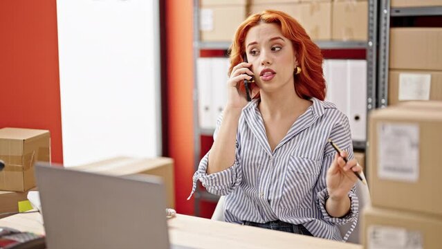 Young redhead woman ecommerce business worker using laptop talking on smartphone at office