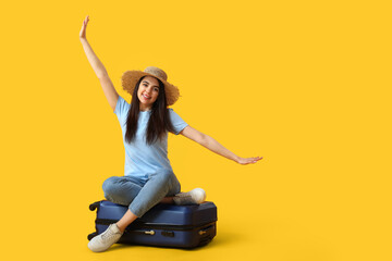 Young woman with suitcase sitting on yellow background