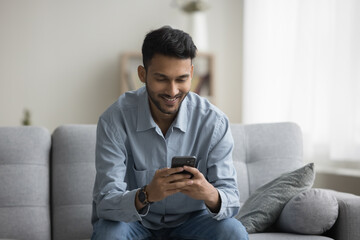 Happy attractive young Indian man using Internet technology at home, typing on cellphone, chatting online with service, application, sitting on sofa in city apartment, smiling, laughing