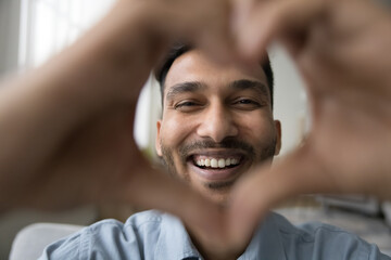 Cheerful attractive young Indian man smiling at camera through hand heart frame. Happy joyful attractive guy showing romantic symbol of love, Valentines day. Indoor male portrait