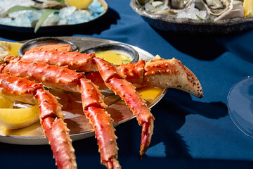 Closeup view of an elegant seafood assortment with prepared crab phalanges, lemon slices, sauces, and fresh oysters on a metal dish, presented on a blue tablecloth, single color blue backdrop.