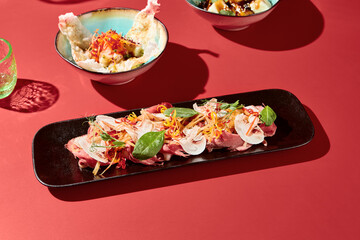 Meat slices, veggies, mushrooms on a black plate; an elegant Asian appetizer. Set against a dark red velvet backdrop with striking shadows, capturing modern minimalist aesthetics