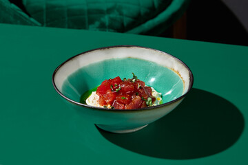 Tuna tartare with stracciatella, placed in a ceramic bowl, served in an Asian restaurant. Stark shadows adding depth, captured horizontally with a view of a green chair in the background