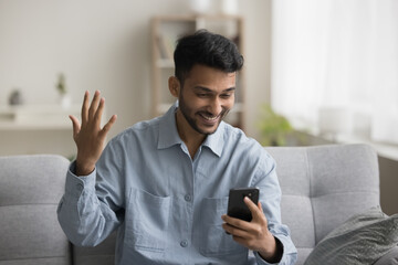 Cheerful excited young Indian freelance business man holding cellphone, looking at screen, making...