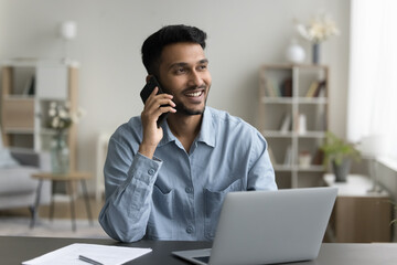 Cheerful young Indian employee guy making job call from home office workplace, speaking on mobile phone, enjoying conversation, talk, communication, looking away, smiling, laughing