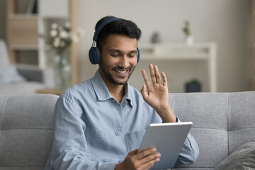 Cheerful young Indian student guy in wireless headphones enjoying online conversation, waving hand hello at tablet webcam, smiling, laughing, enjoying Internet technology connection