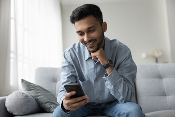 Cheerful handsome young adult Indian guy holding mobile phone, enjoying leisure with gadget, having fun, chatting, talking on online video call, reading, watching Internet content