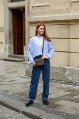 a red-haired girl in a blue jeans and a sweater poses outside with a small leather handbag