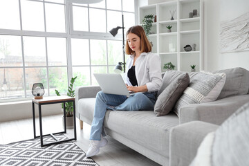 Pretty young woman sitting on grey sofa and using modern laptop in light living room