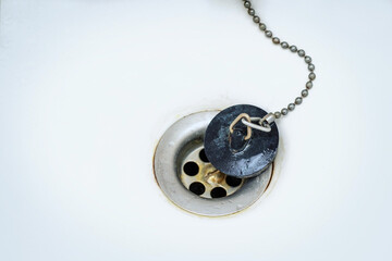 Old black rubber plug with metal chain on a rusty drain in a white wash basin or bathtub, copy...