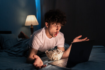 Amazed guy eating popcorn and watching horror movie on laptop while lying in bed at night