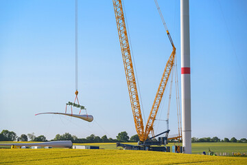 Giant crane lifting a wind turbine blade to install it onto the tower, heavy industry construction site, concept for electricity, renewable energy and power, blue sky, copy space - 611904280