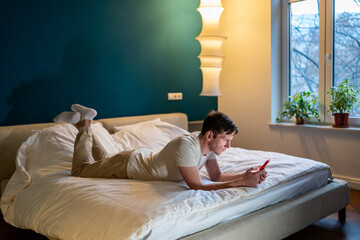Man indoors at home lying on bed, using mobile phone chatting. Brunette male flirting in social...