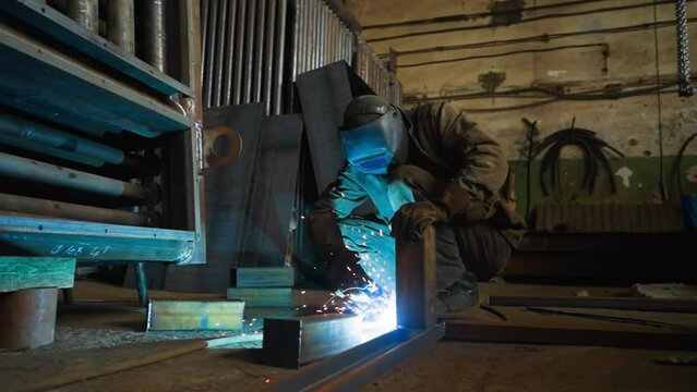 Welder touches connected details of beam frame with metal electrode on floor in plant workshop slow motion. Employee works with welding tool at plant