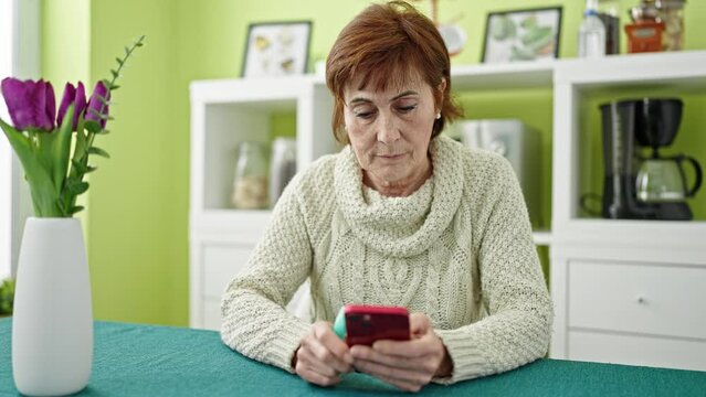Mature hispanic woman holding vape inhaler sitting on table using smartphone at dinning room