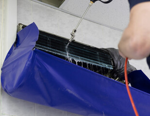 A man's hand sprays pressurized water onto an air conditioner grille. A man washes away dirt and...