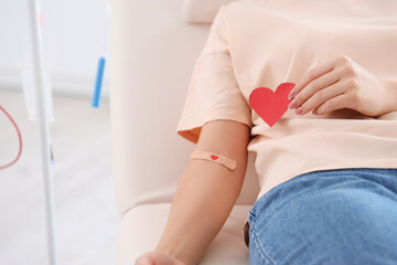 Female blood donor with applied medical patch and paper heart in clinic, closeup