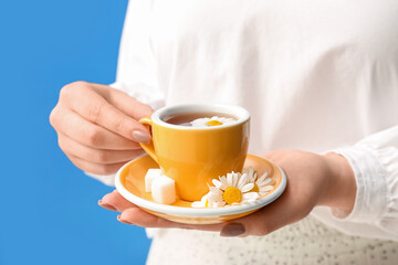 Beautiful young woman with cup of chamomile tea and flowers near blue wall