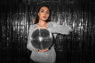 Fashionable beautiful woman in sparkling dress with disco ball posing against silver tinsel